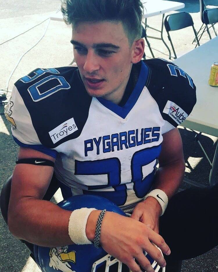 A young, smooth-faced jock sits outdoors wearing a full blue and white football kit while holding his helmet.