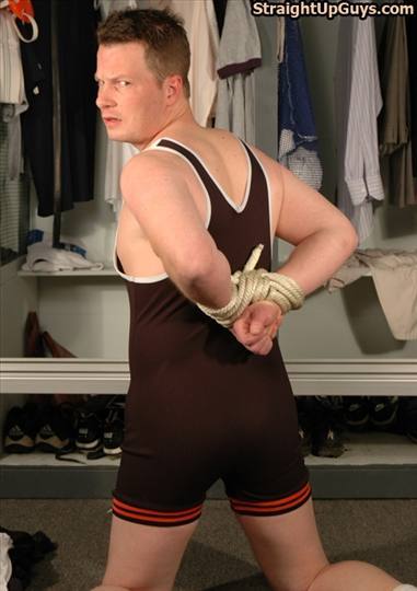 A man in a wrestling singlet has his wrists bound behind his back with rope while kneeling in a closet.