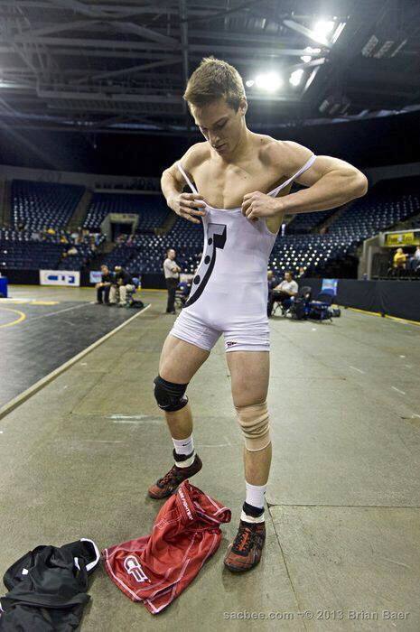 A muscular young man pulls on a white spandex wrestling singlet while standing in an indoor sports arena.