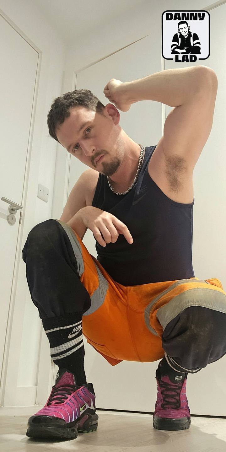 A bearded twunk in hi-vis construction trousers and a black tank top crouches while displaying his hairy armpit.