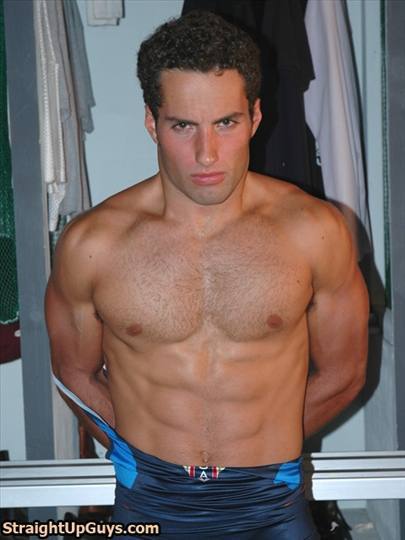 A muscular jock with a hairy chest stands in a locker room while pulling down his blue wrestling singlet.
