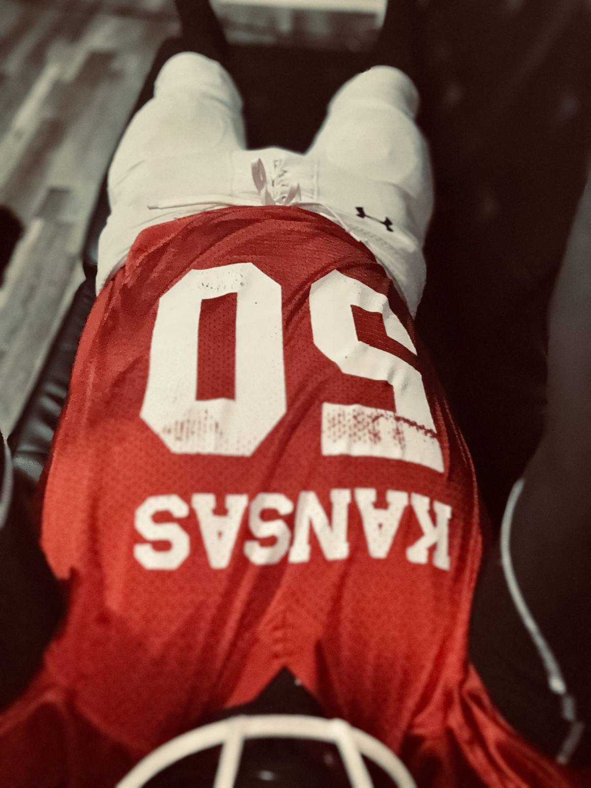 A person wearing a red Kansas football jersey and white padded football pants lies on a dark surface with their helmet visible at the bottom of the frame.