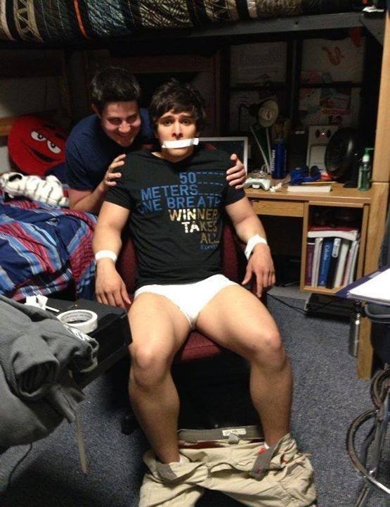 A young man is taped to a chair in a dorm room with duct tape over his mouth and around his wrists while wearing white briefs with his pants pulled down.