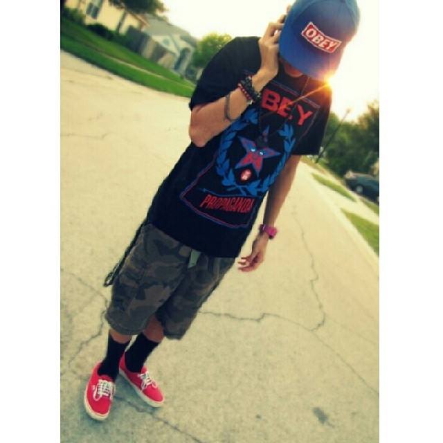 A young man in streetwear, featuring a blue cap, graphic tee, camo shorts, and red sneakers, stands on a residential street.