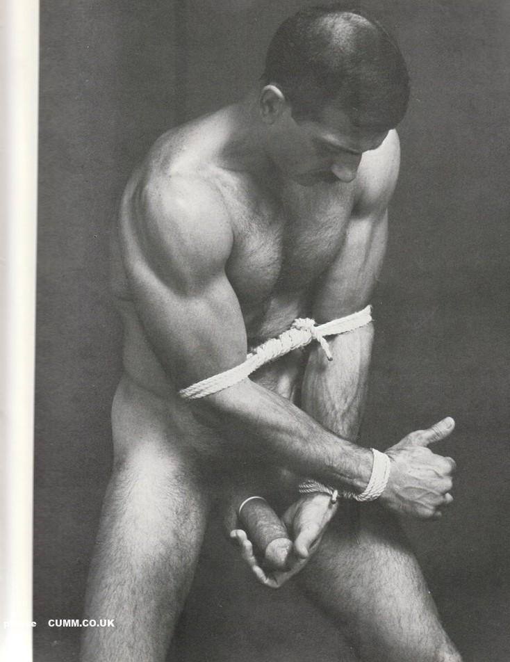 A muscular man with a hairy chest stands nude, bound by white rope around his biceps and wrists.