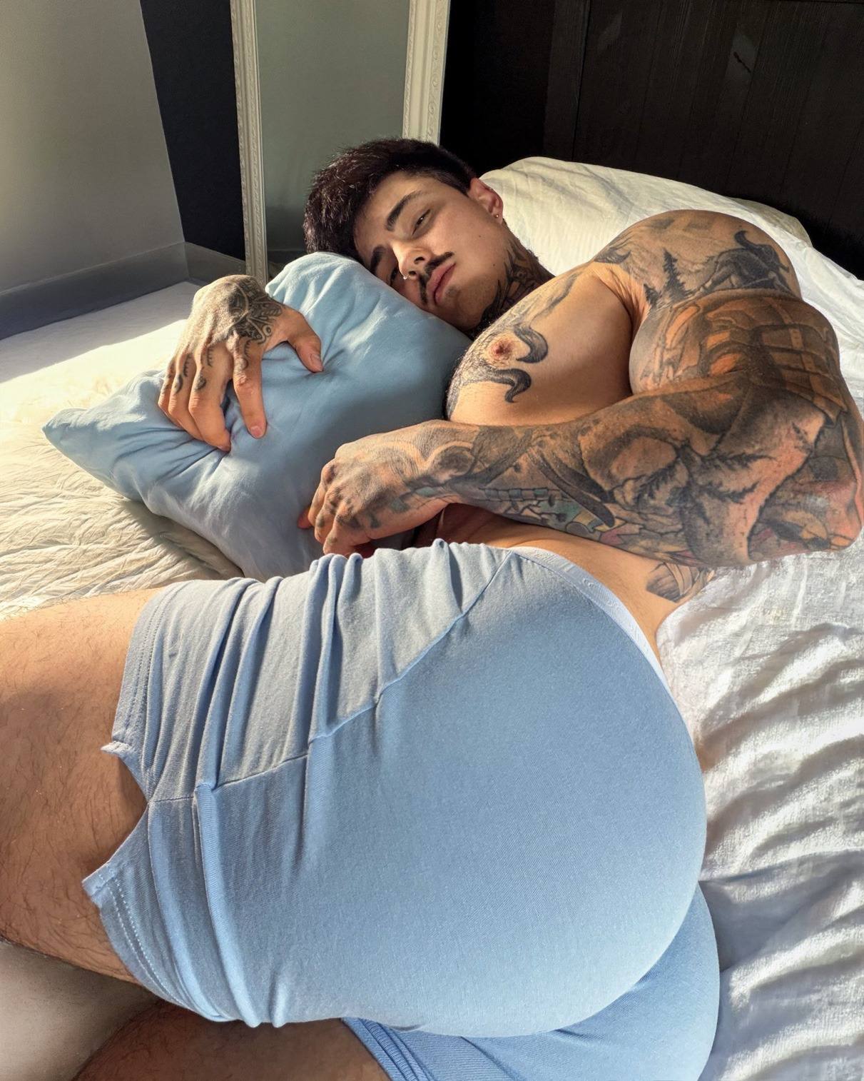 A muscular man with extensive tattoos lies on a bed in light blue boxer briefs while holding a matching pillow.
