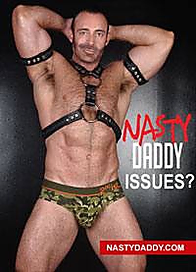 A muscular mature man poses wearing a black studded leather chest harness, matching armbands, and camouflage briefs against a dark background.