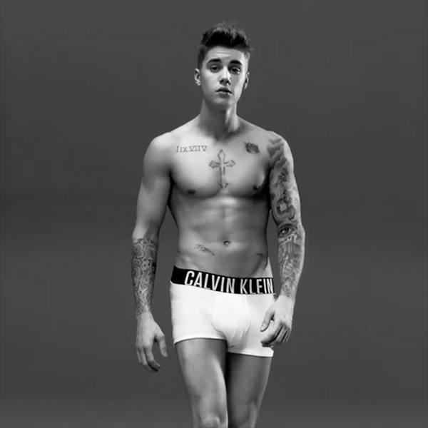 A lean, tattooed young man poses in white Calvin Klein boxer briefs against a plain studio background.