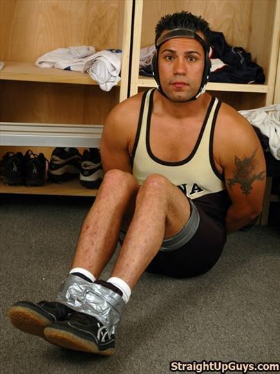 A muscular man in a wrestling singlet and headgear sits on a locker room floor with his ankles bound by duct tape and hands restrained behind his back.