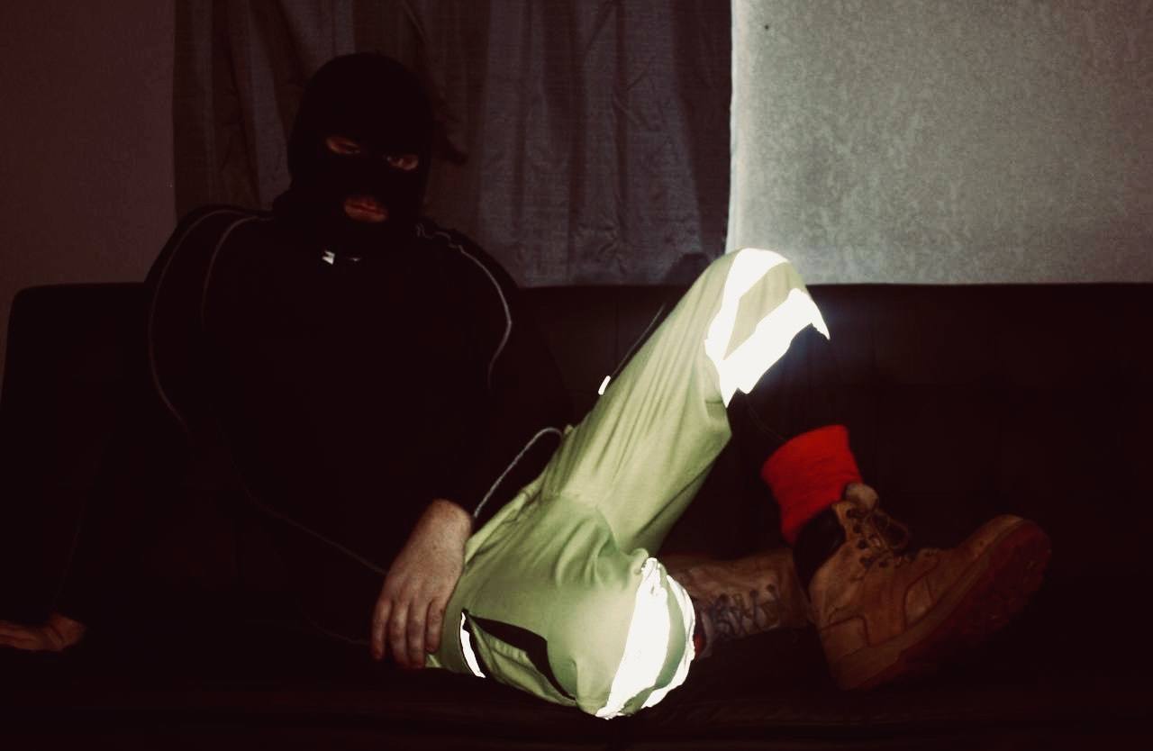 A man wearing a black balaclava and high-visibility construction trousers sits on a dark couch.