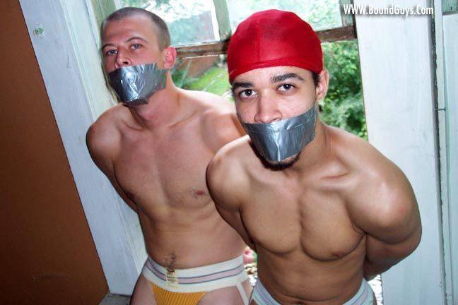 Two shirtless young men wearing jockstraps stand bound behind their backs with silver duct tape gags over their mouths.