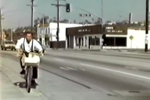 A man wearing a white shirt and suspenders rides a bicycle along a public city street.