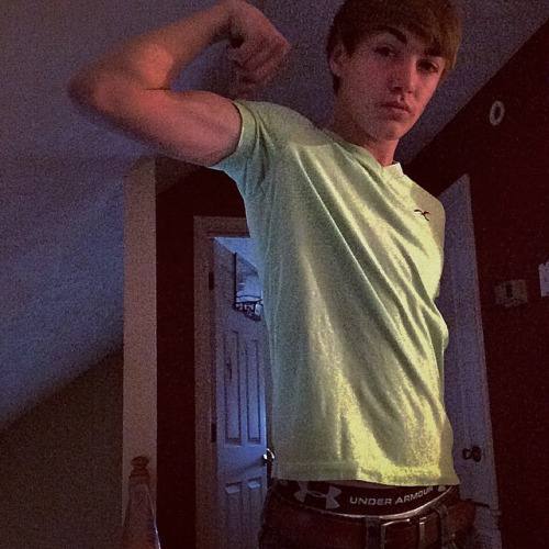 A young man flexes his bicep in a bedroom while wearing a lime green t-shirt and showing his Under Armour waistband.