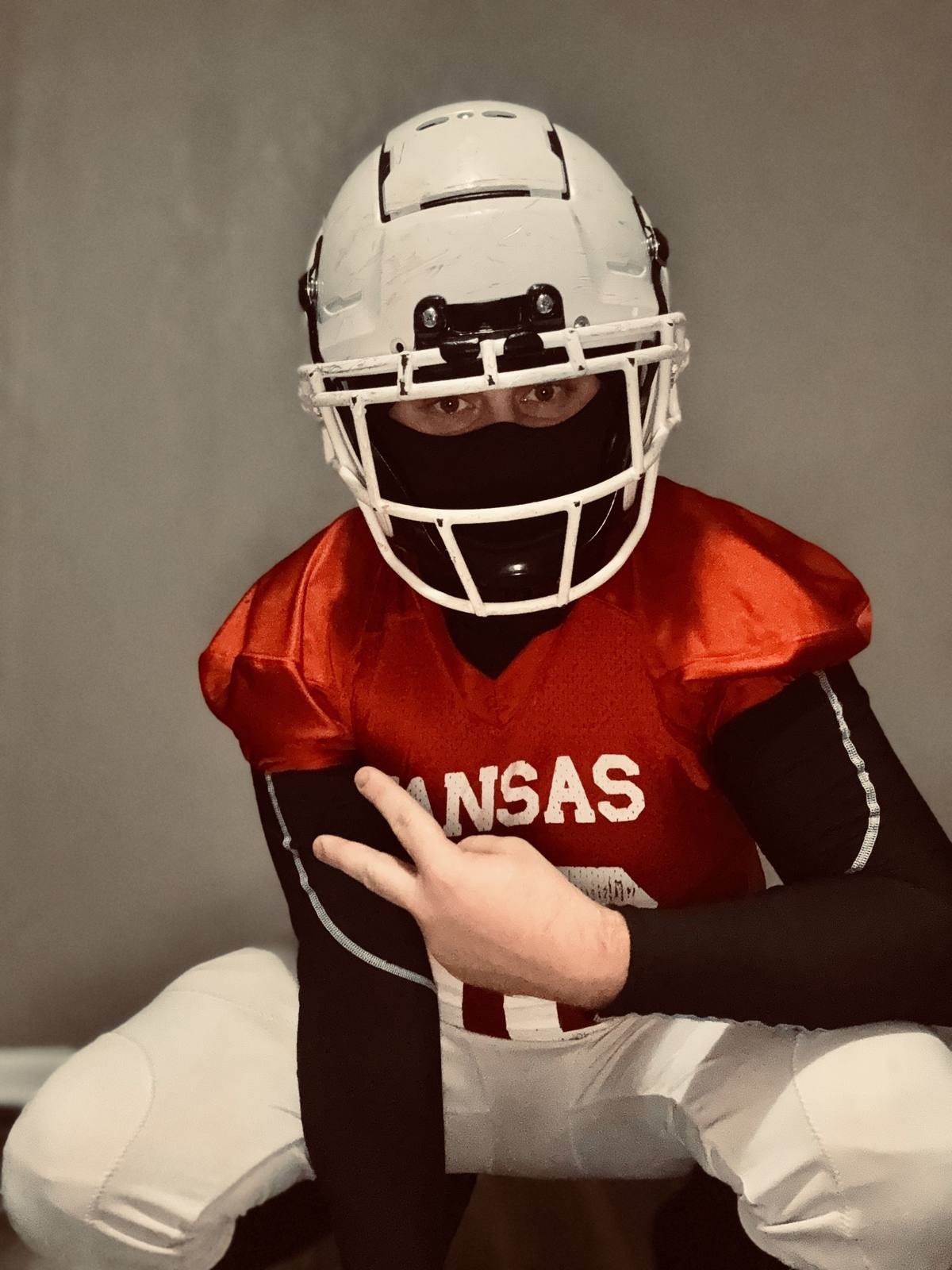 A man wearing a full American football kit, including a helmet and black balaclava, squats while posing for a photo.