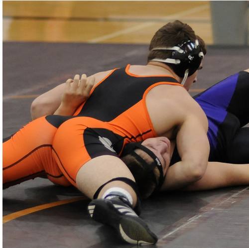 Two male wrestlers wearing spandex singlets and headgear grapple on a mat inside a gymnasium.