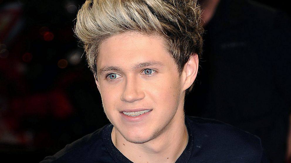 A close-up portrait of a smooth young man with styled blonde hair and blue eyes wearing braces and a dark shirt.