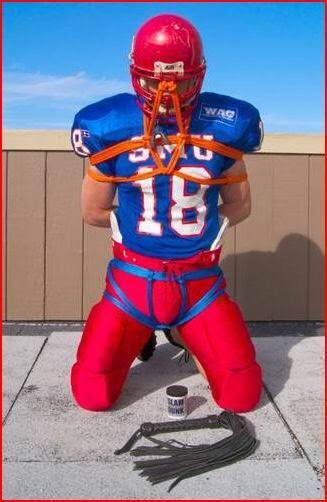 A muscular man in a full football uniform is kneeling outdoors while bound with orange rope, with a leather flogger on the ground in front of him.