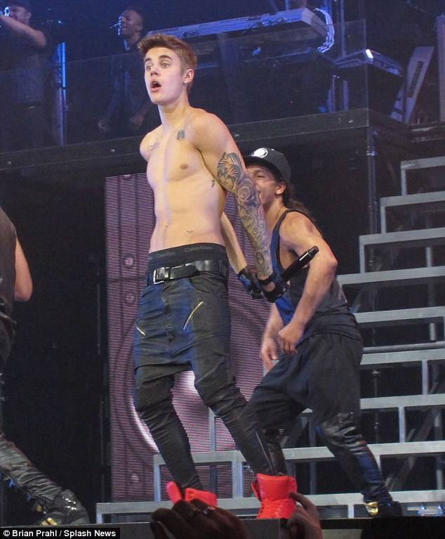 A lean, shirtless young man with a tattooed arm wears sagging black jeans and boxer briefs while performing on a stage.