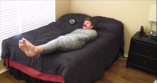 A young man lies on a bedroom bed, tightly cocooned in silver duct tape from shoulders to feet with a red gag in his mouth.