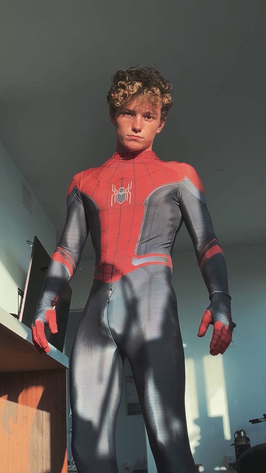 A young man stands indoors wearing a form-fitting Spider-Man costume with a front zipper.
