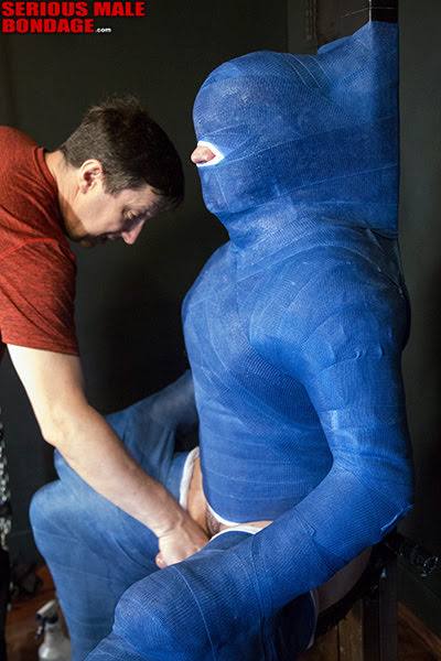 A mature man interacts with a subject completely encased in a blue fiberglass body cast within a dungeon setting.