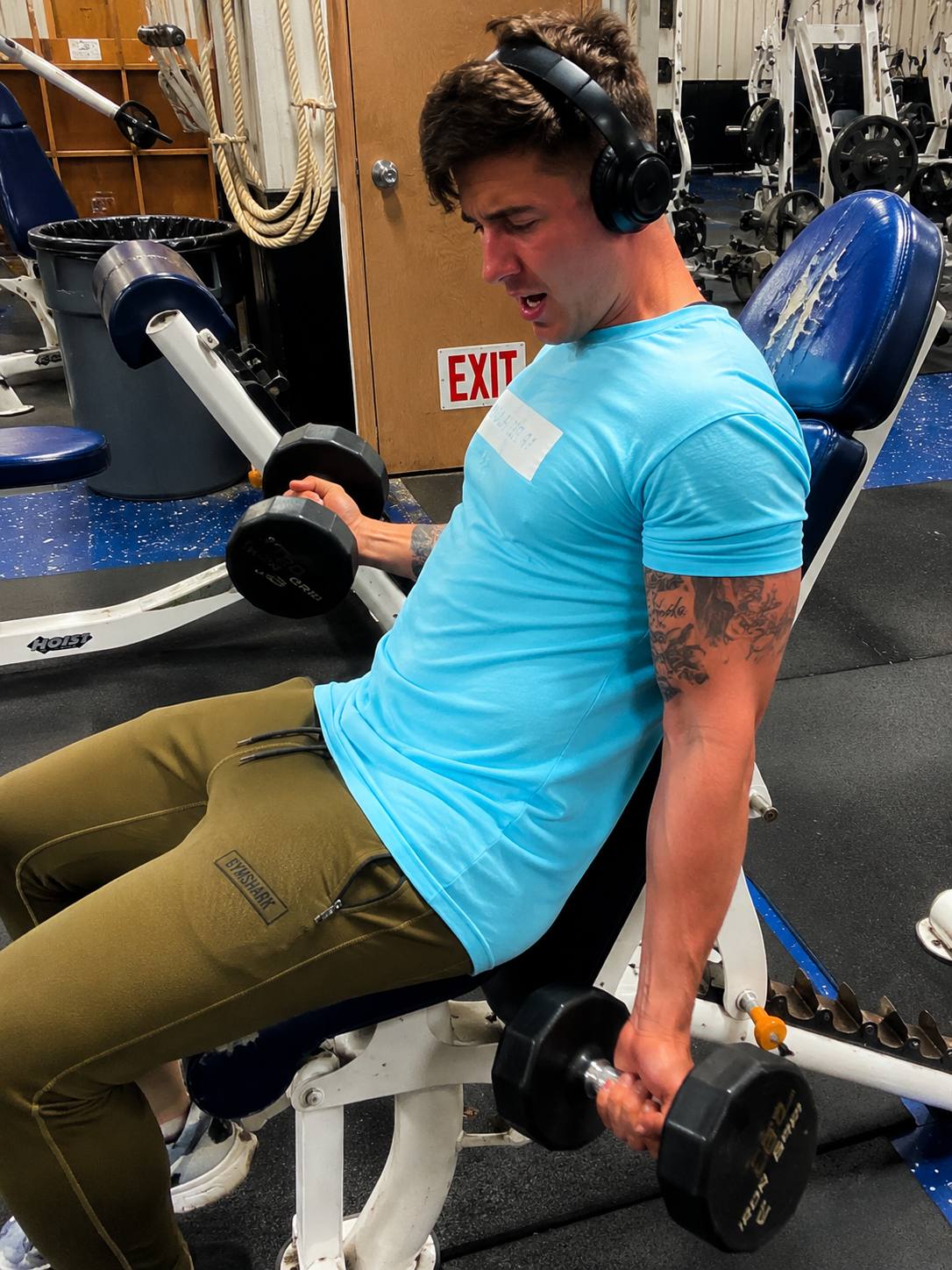A muscular man in a tight blue shirt and olive sweatpants performs dumbbell curls in a gym.