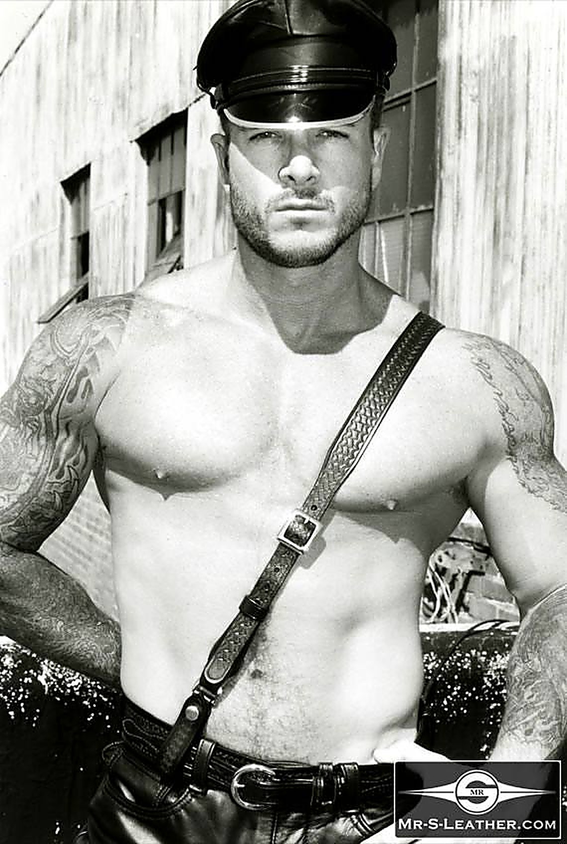 A shirtless muscular tattooed man wearing a leather muir cap and a leather crossbody strap poses outdoors.