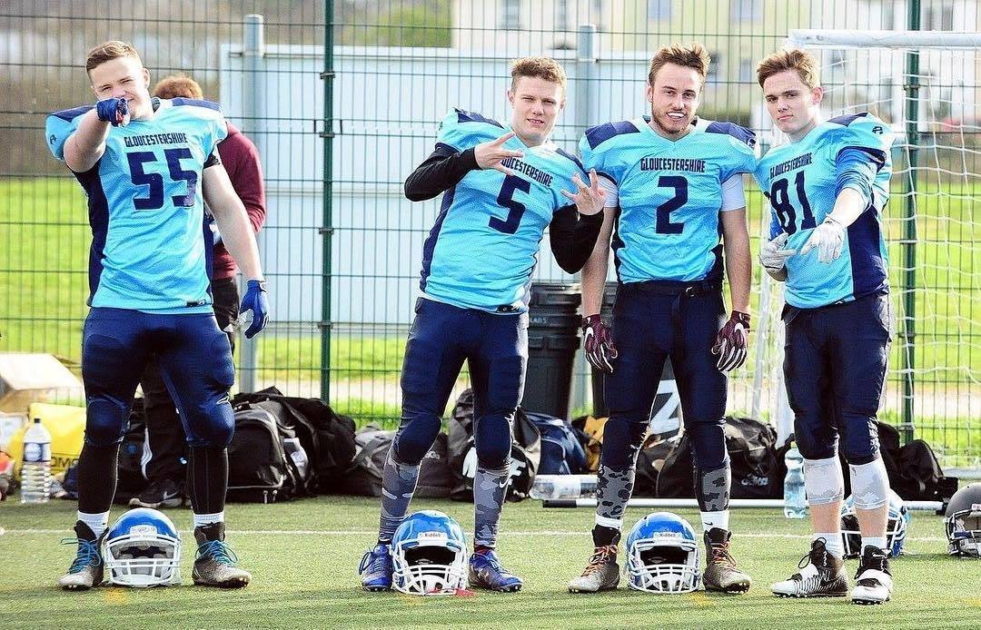 Four young men pose on an outdoor sports field wearing light blue and dark blue American football kits.