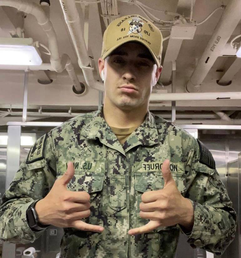 A young man in a US Navy digital camouflage uniform and tan cap poses inside a ship or industrial facility.