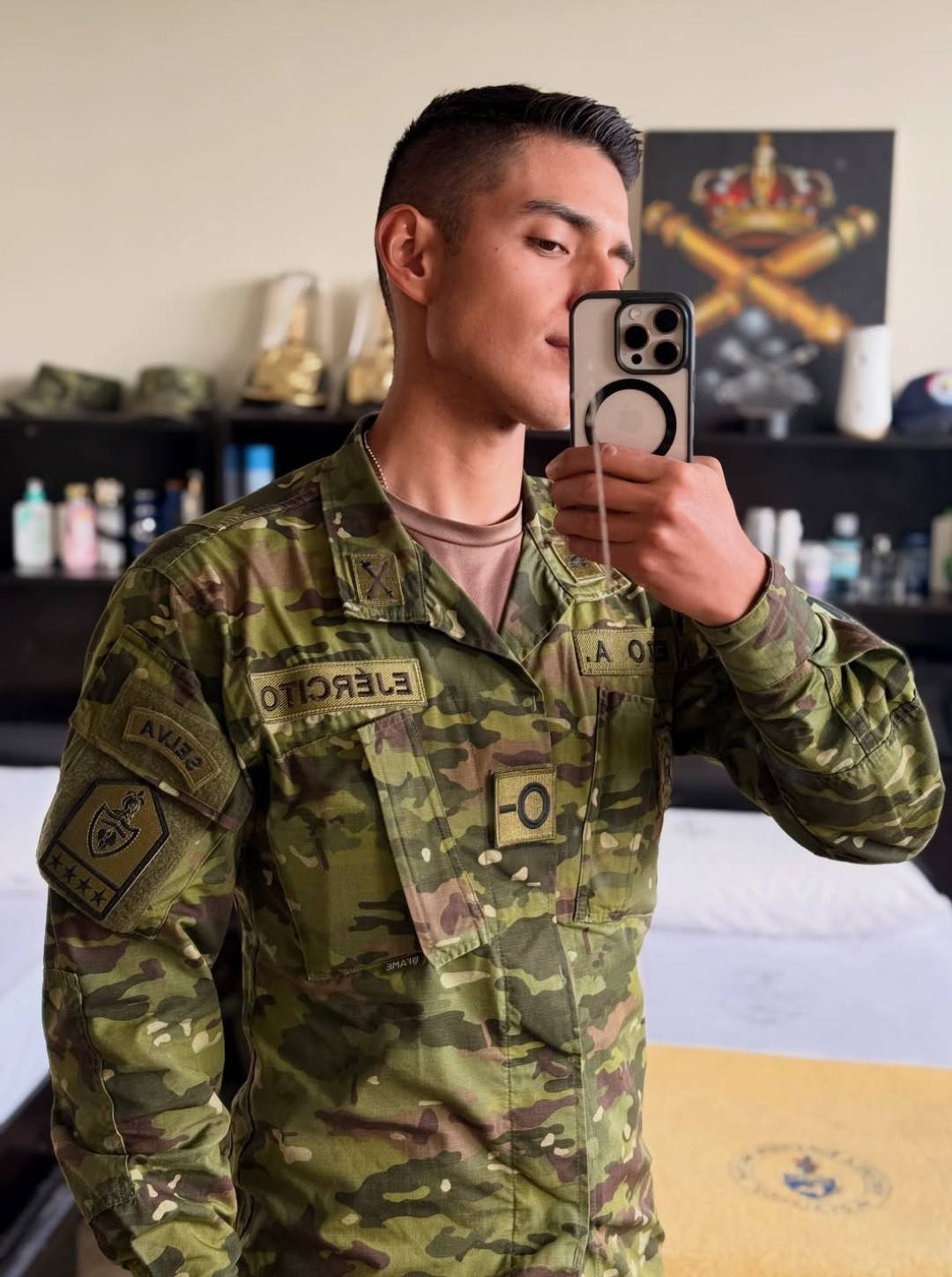 A young man with a short fade haircut takes a mirror selfie while wearing military camouflage fatigues.
