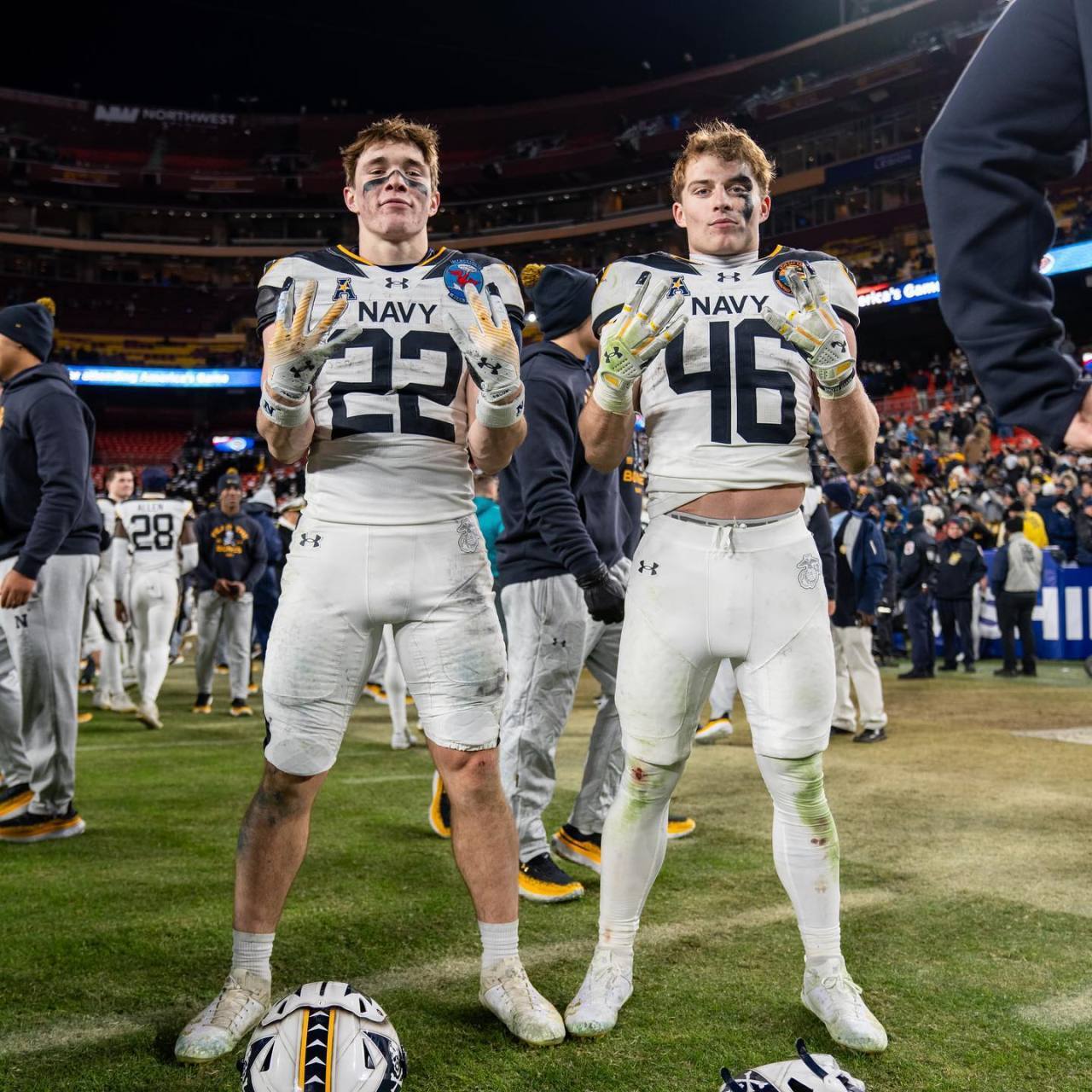 Two athletic young men in Navy football uniforms pose on a stadium field wearing tight compression pants and gloves.