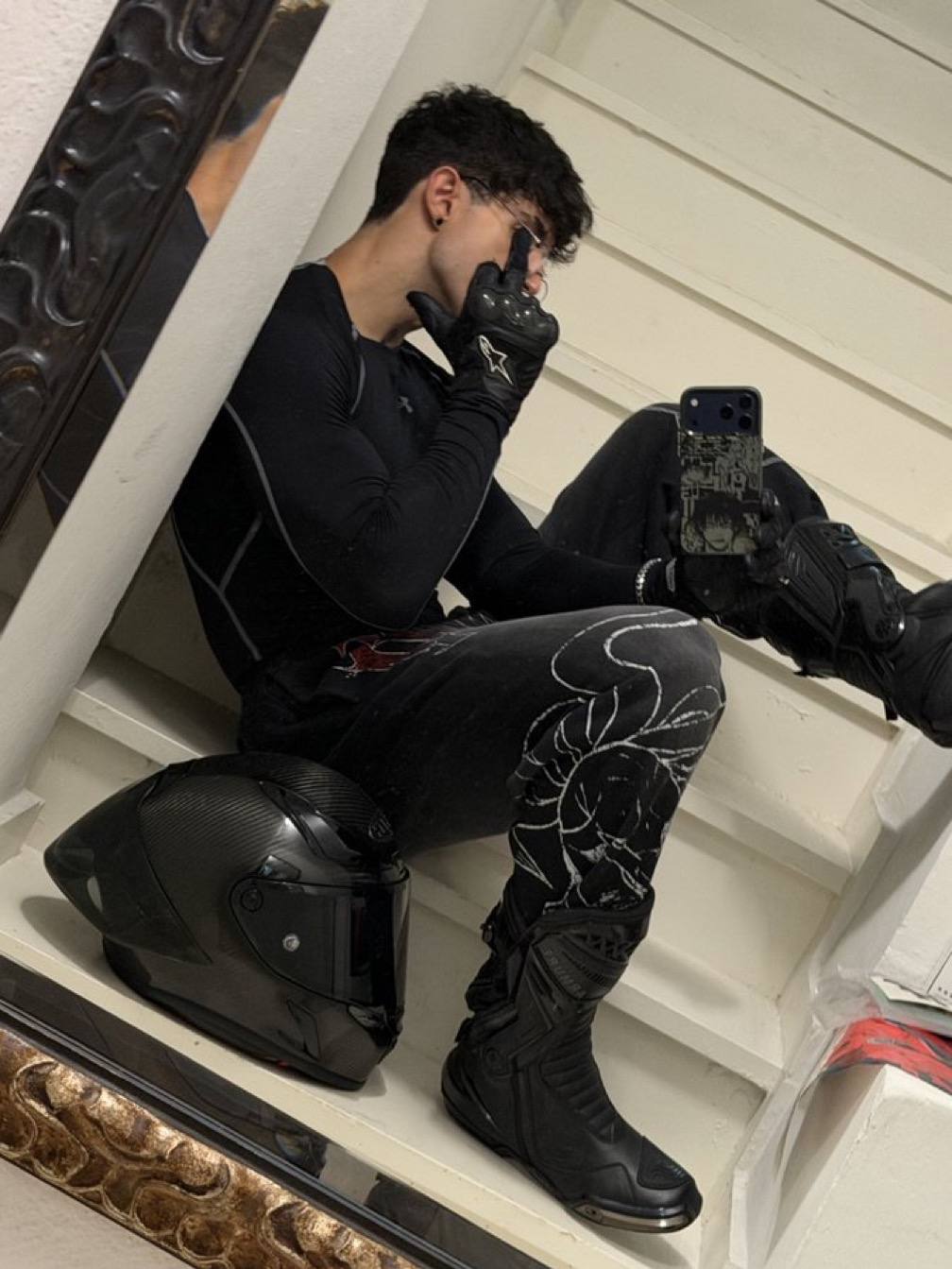A young man in a black compression top, motorcycle boots, and gloves poses for a mirror selfie on a staircase next to his helmet.