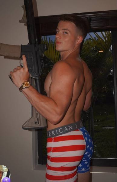 A muscular young man with a buzzcut poses shirtless in patriotic American flag boxer briefs while holding a large tactical rifle.