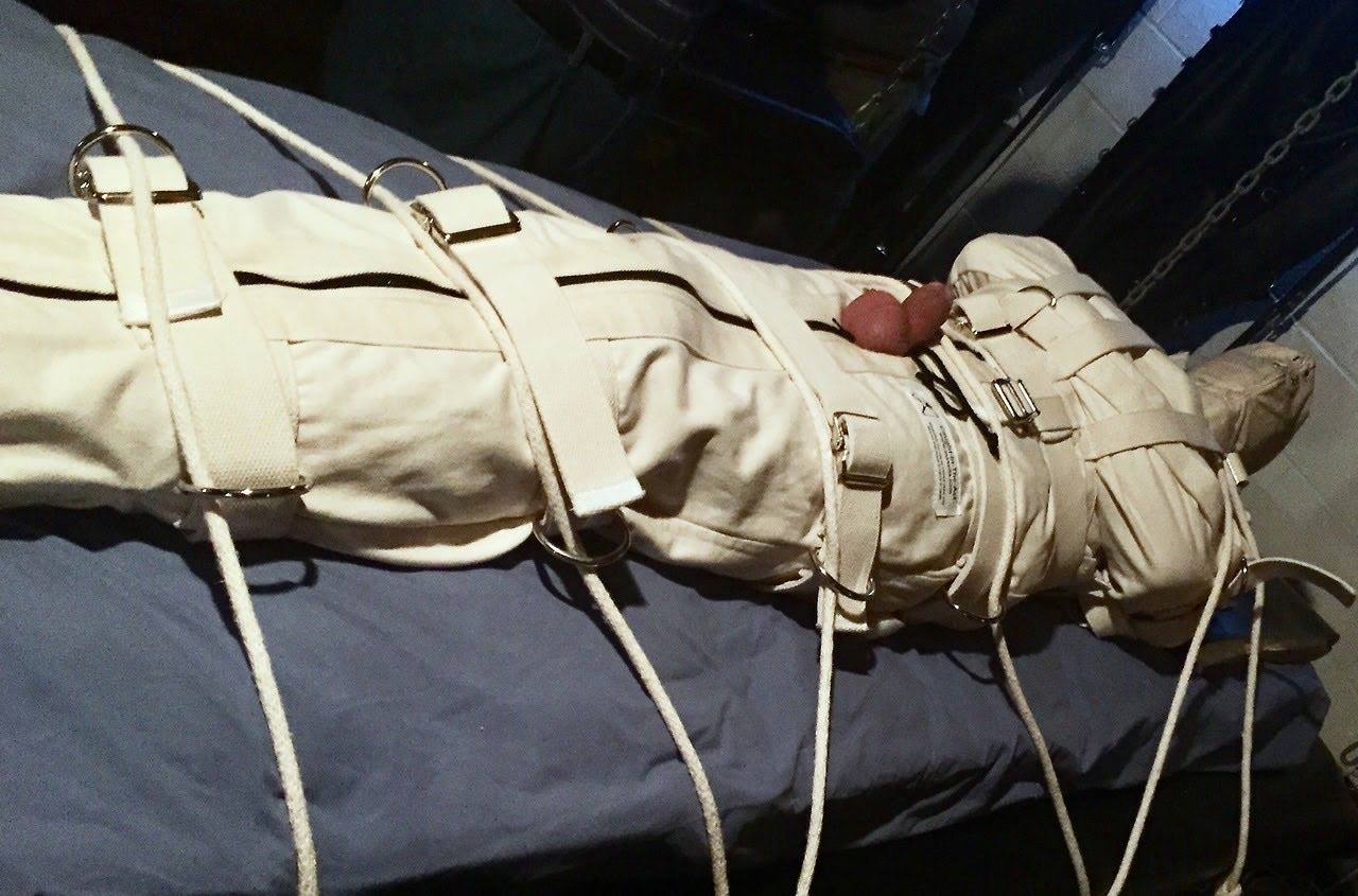 A man is completely immobilized in a heavy white canvas bondage suit with his genitals exposed, secured to a surface with ropes in a dungeon setting.