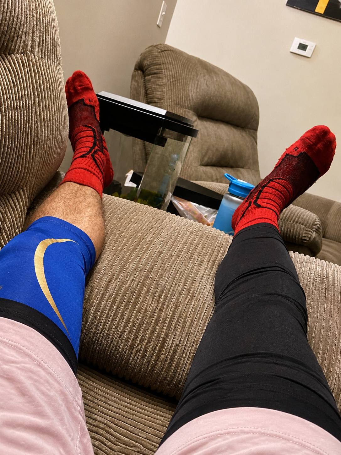 A POV shot of a man relaxing on a couch wearing red athletic socks and mismatched compression gear on his hairy legs.