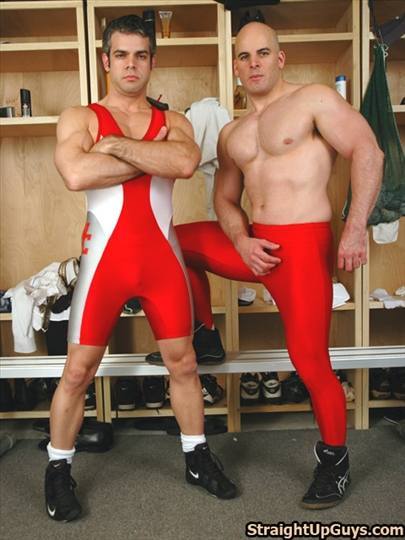 Two muscular men in red and white wrestling gear pose together in a locker room setting.