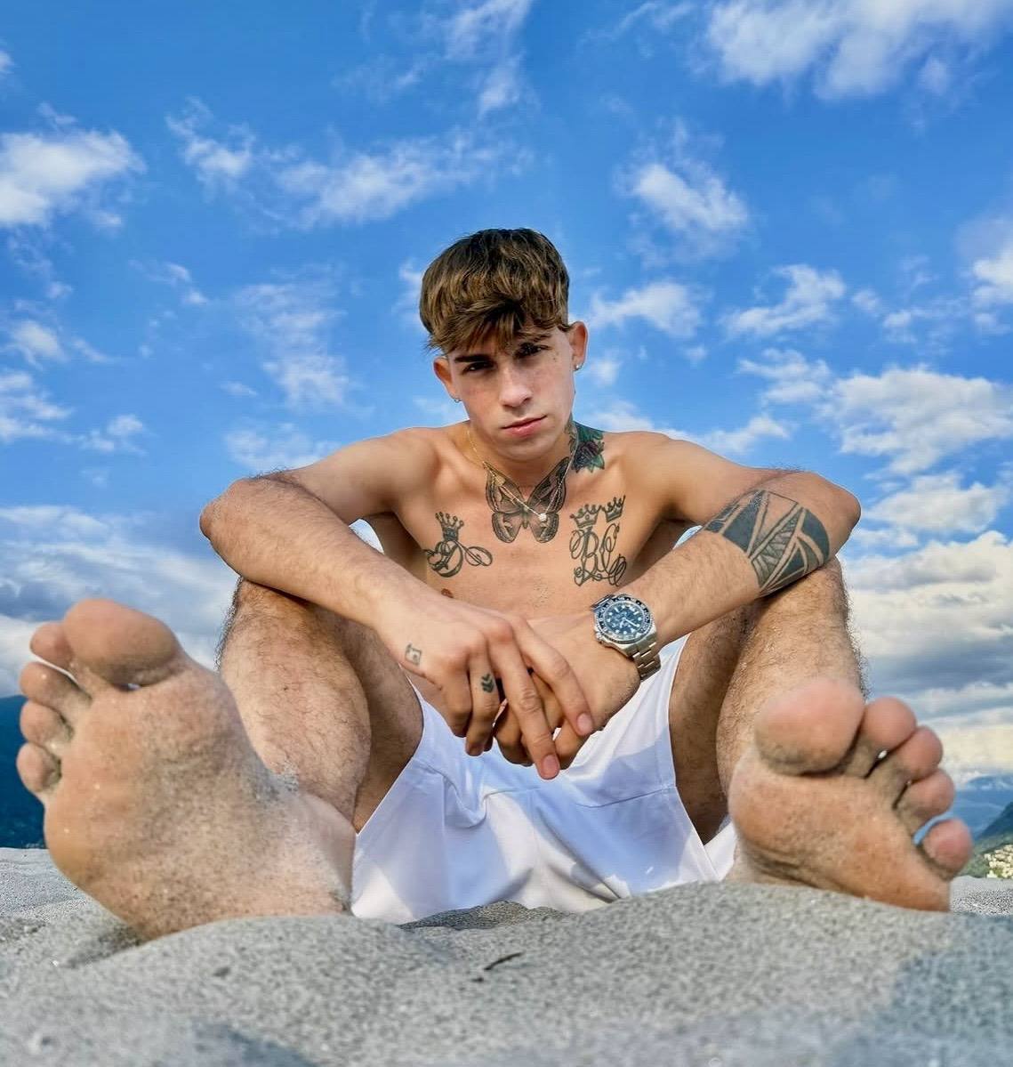 A tattooed young man sits on a sandy beach with his bare, sand-covered soles prominently displayed toward the camera.