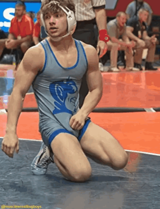 A muscular young wrestler wearing a grey singlet and headgear kneels on the mat while adjusting his crotch.