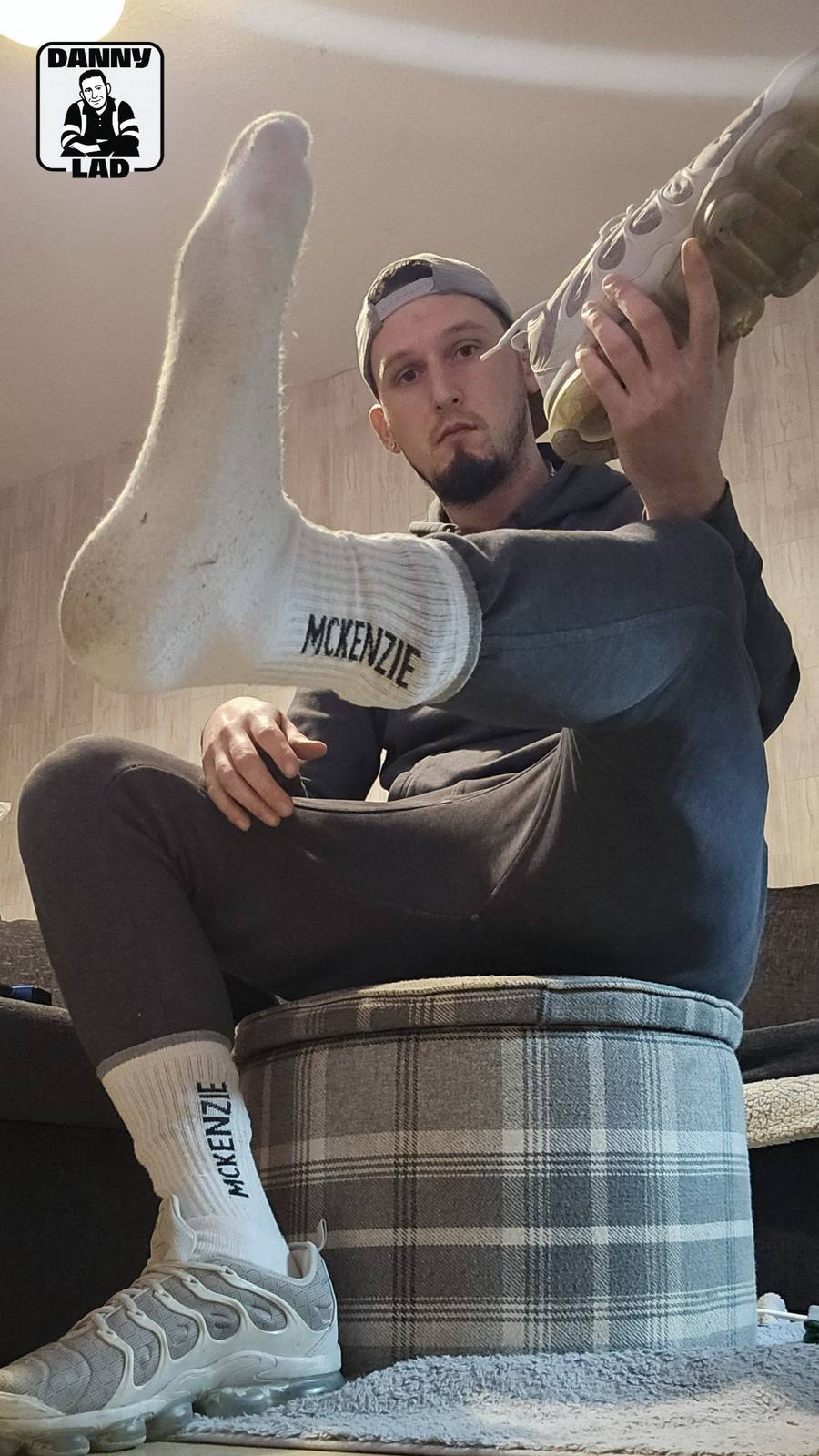 A man in a grey tracksuit and backwards cap poses with his feet in white McKenzie crew socks and Nike sneakers.