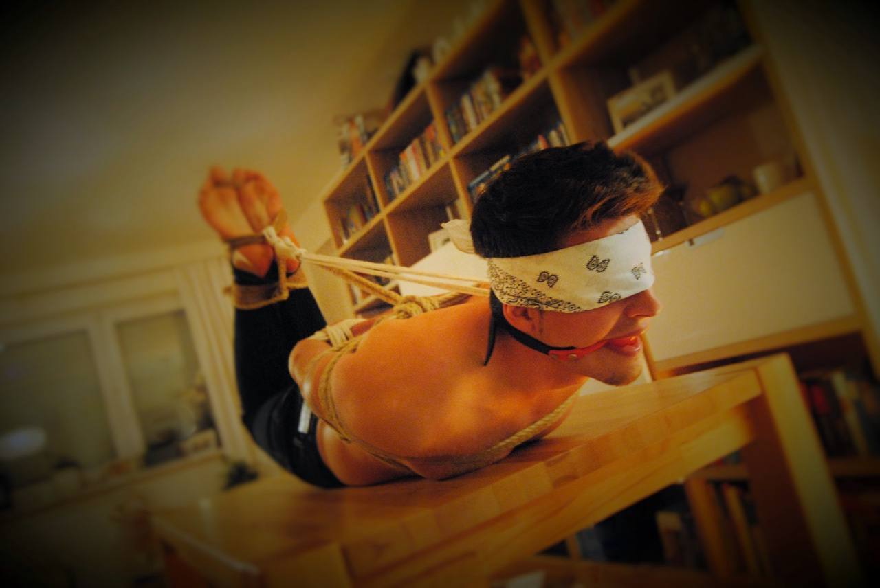A shirtless young man lies prone on a wooden table, restrained in a rope hogtie with a blindfold and ball gag.