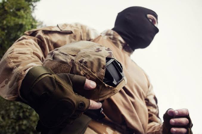 A person dressed in a desert camouflage uniform and a black balaclava holds a tactical helmet in an outdoor setting.