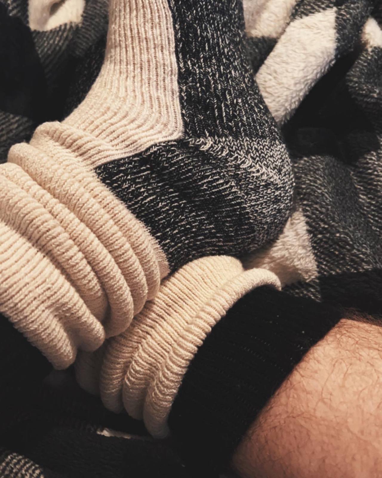 A close-up view of hairy legs wearing thick, cream-colored ribbed slouch socks over dark wool socks on a checkered blanket.