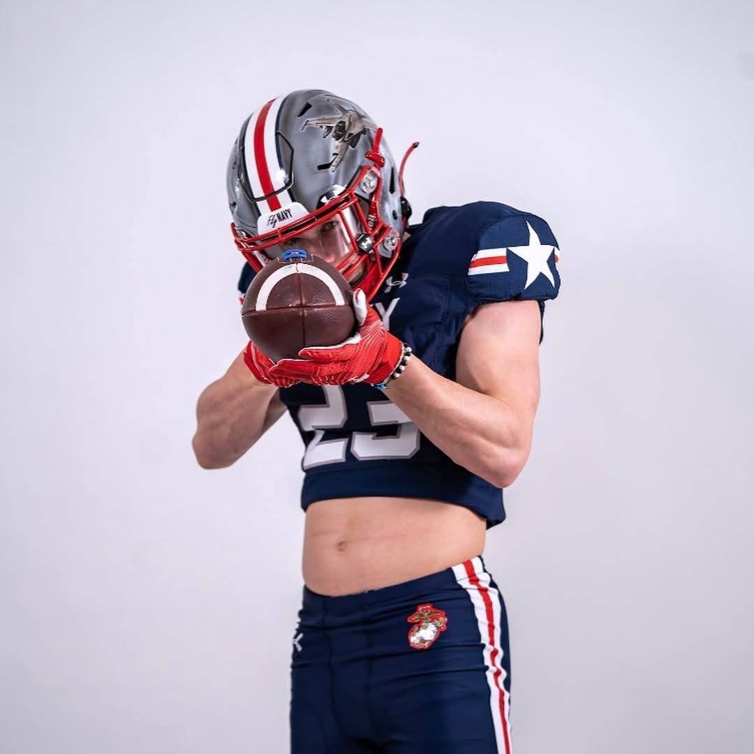 A muscular young man in a full Navy football kit with a cropped jersey poses while holding a football, revealing his toned midriff and abs.