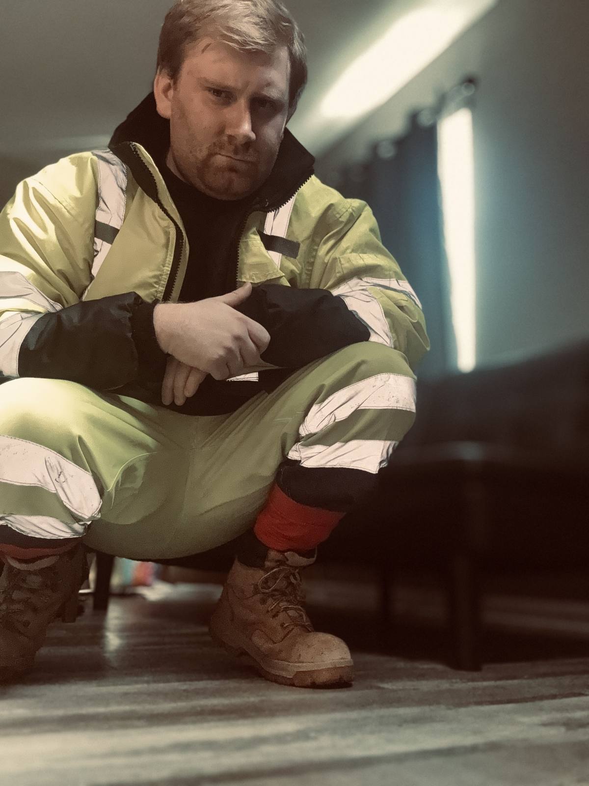 A bearded man wearing a full high-visibility construction uniform and work boots squats in an indoor setting.