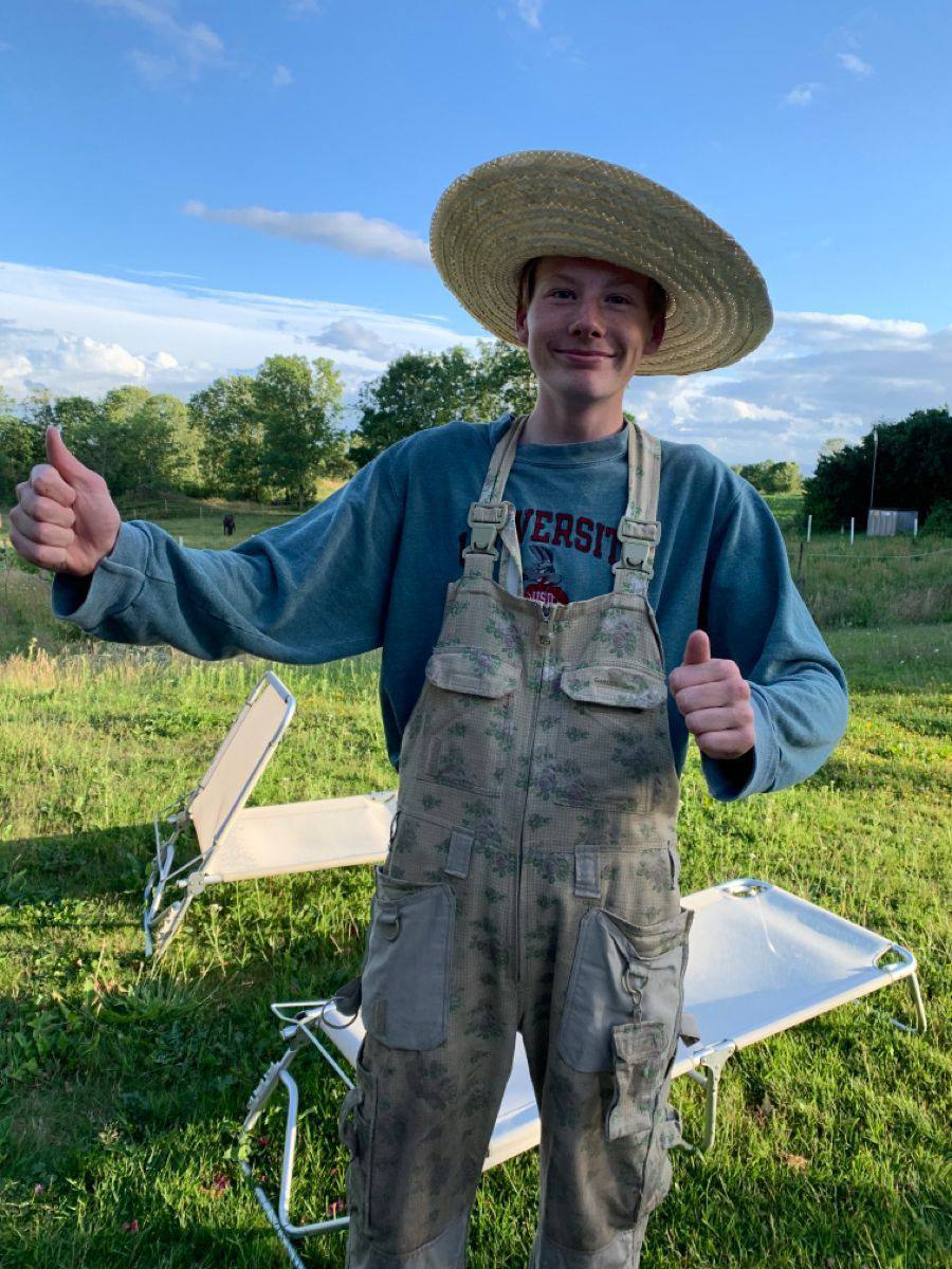 A young man in a straw hat and overalls smiles and gives a thumbs up outdoors.