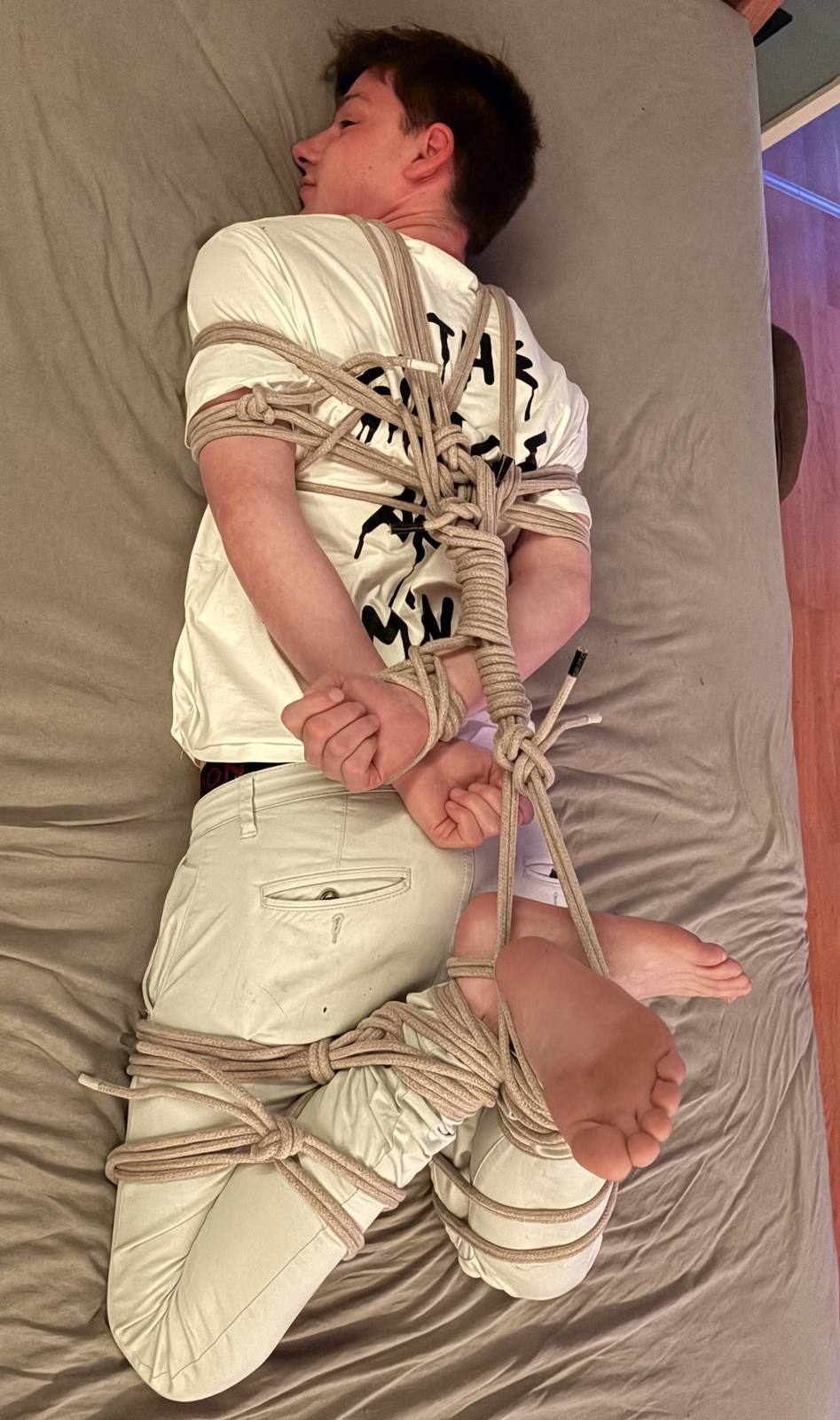 A young man is bound in complex rope bondage, lying on a bed with his hands and legs tied behind him.