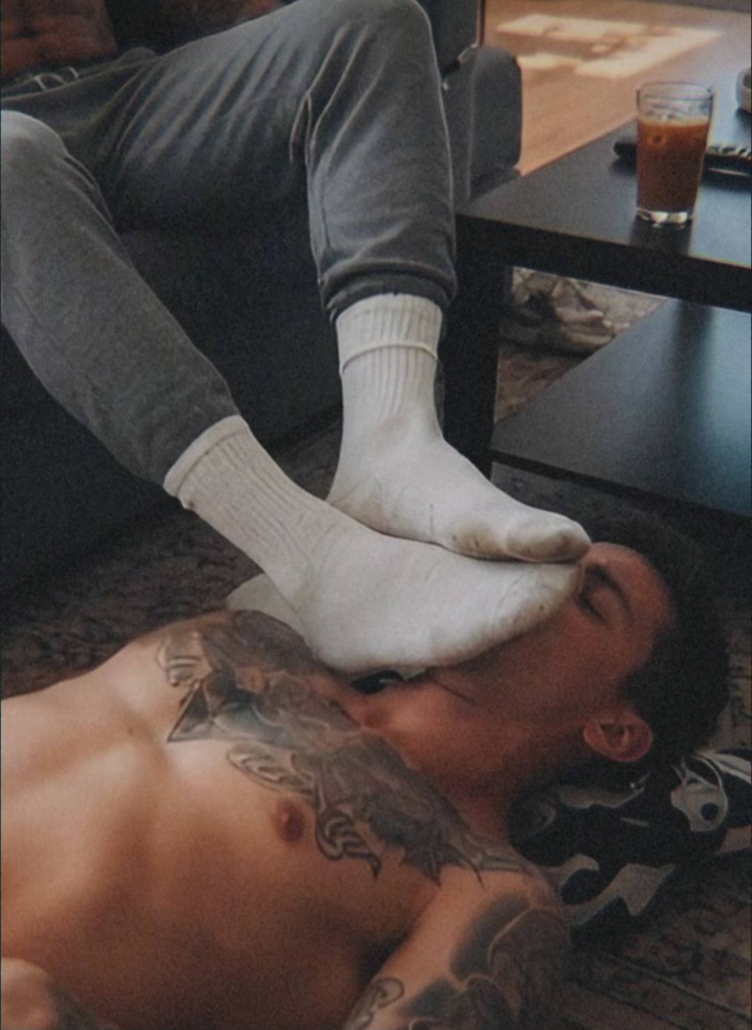 A man lying on the floor has a pair of white tube-socked feet pressed against his face by another person sitting on a sofa.
