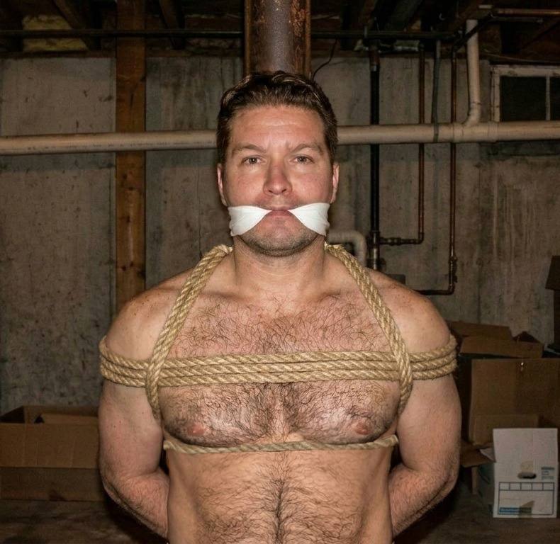 A shirtless muscular man with a hairy chest is gagged and bound with rope to a support beam in a basement setting.