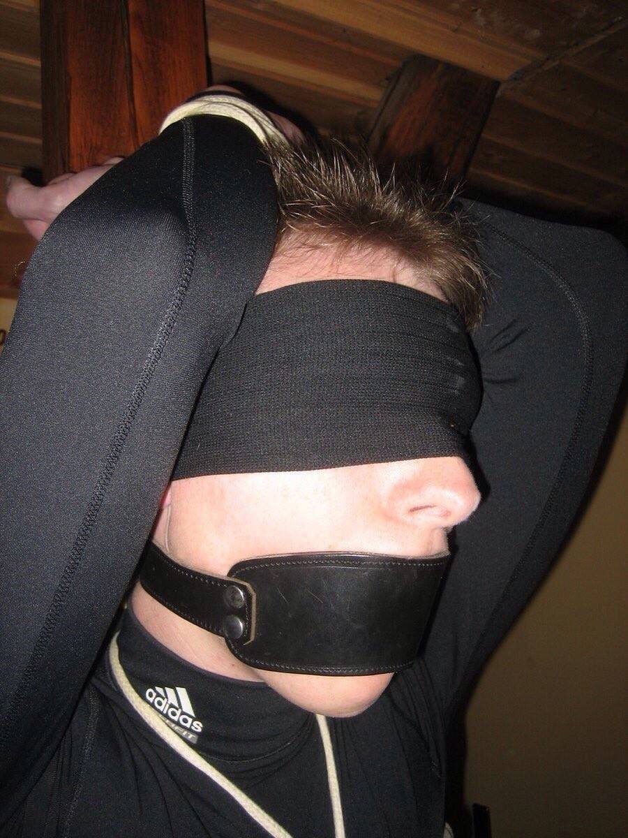 A young submissive man wearing a black elastic blindfold, leather panel gag, and compression shirt has his hands bound overhead.