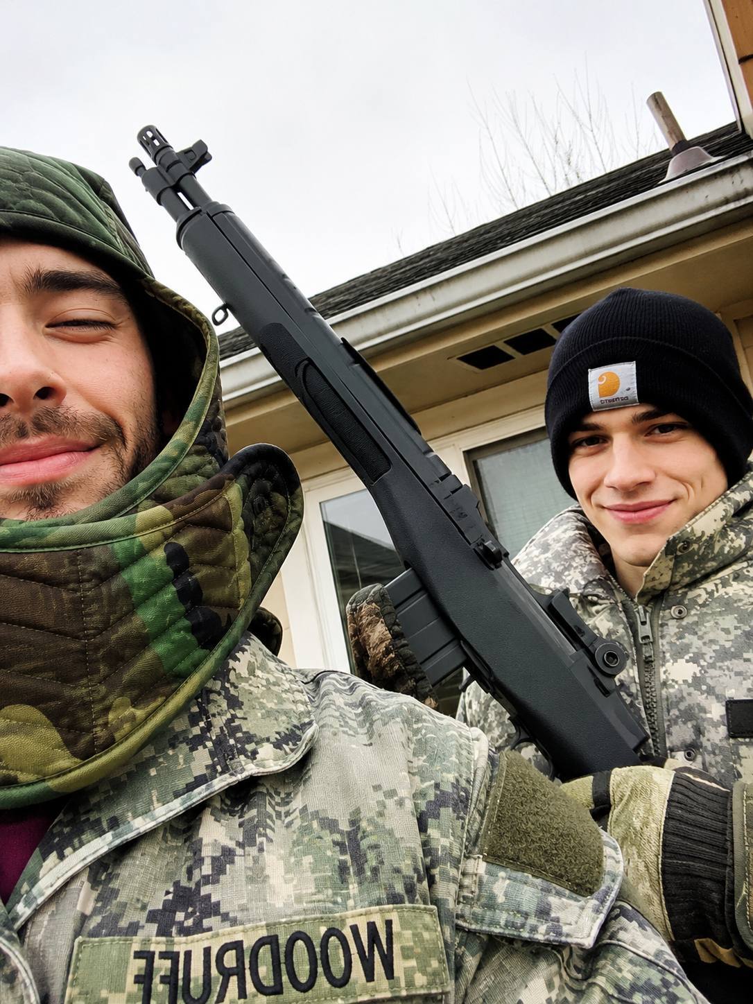 Two young men in military camouflage uniforms pose outdoors while holding a black rifle.